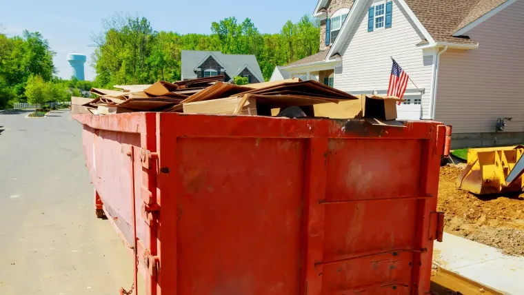 Red construction dumpster filled with debris on a suburban street beside a house