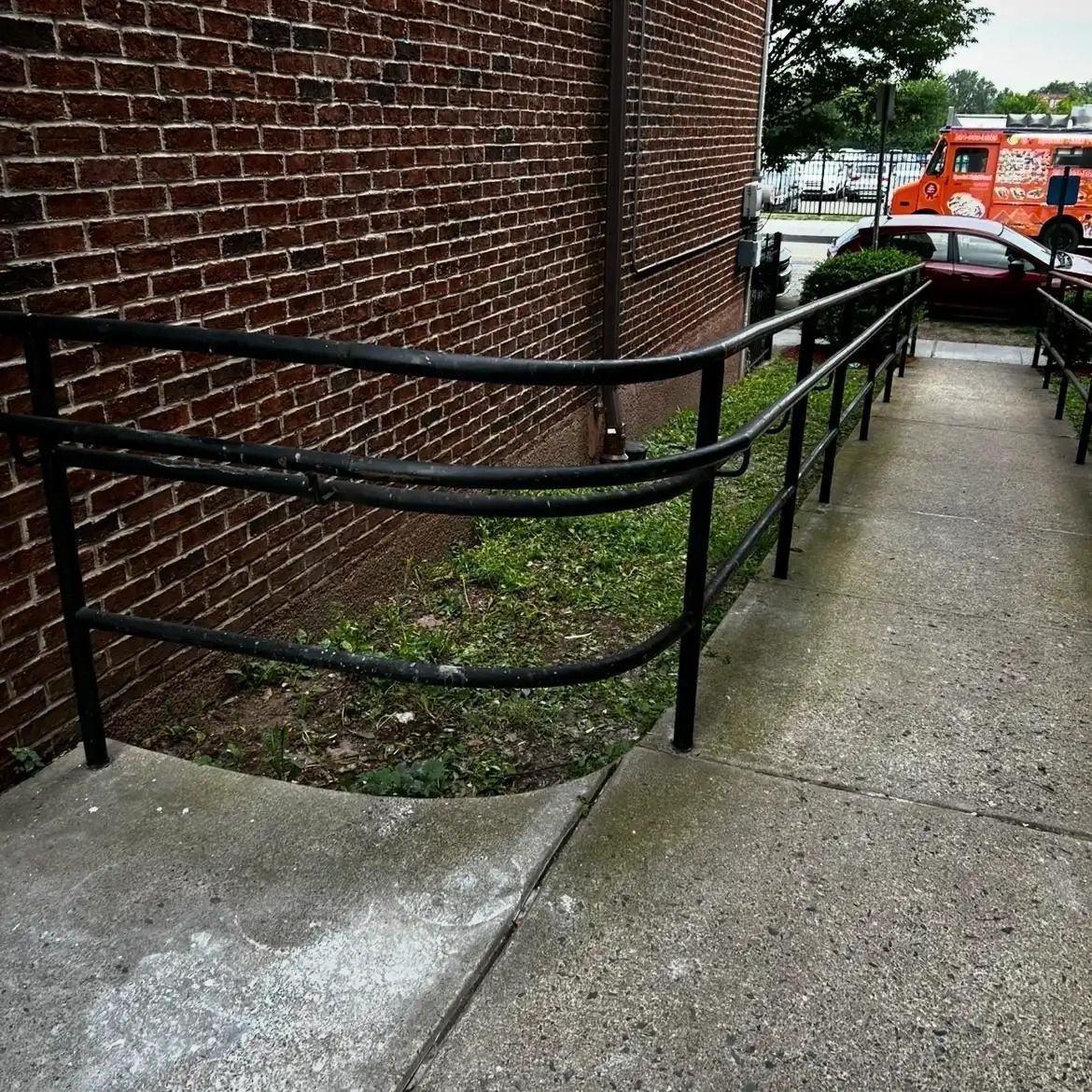 Black handrails along a sloped concrete ramp beside a brick building, with an ambulance in the background