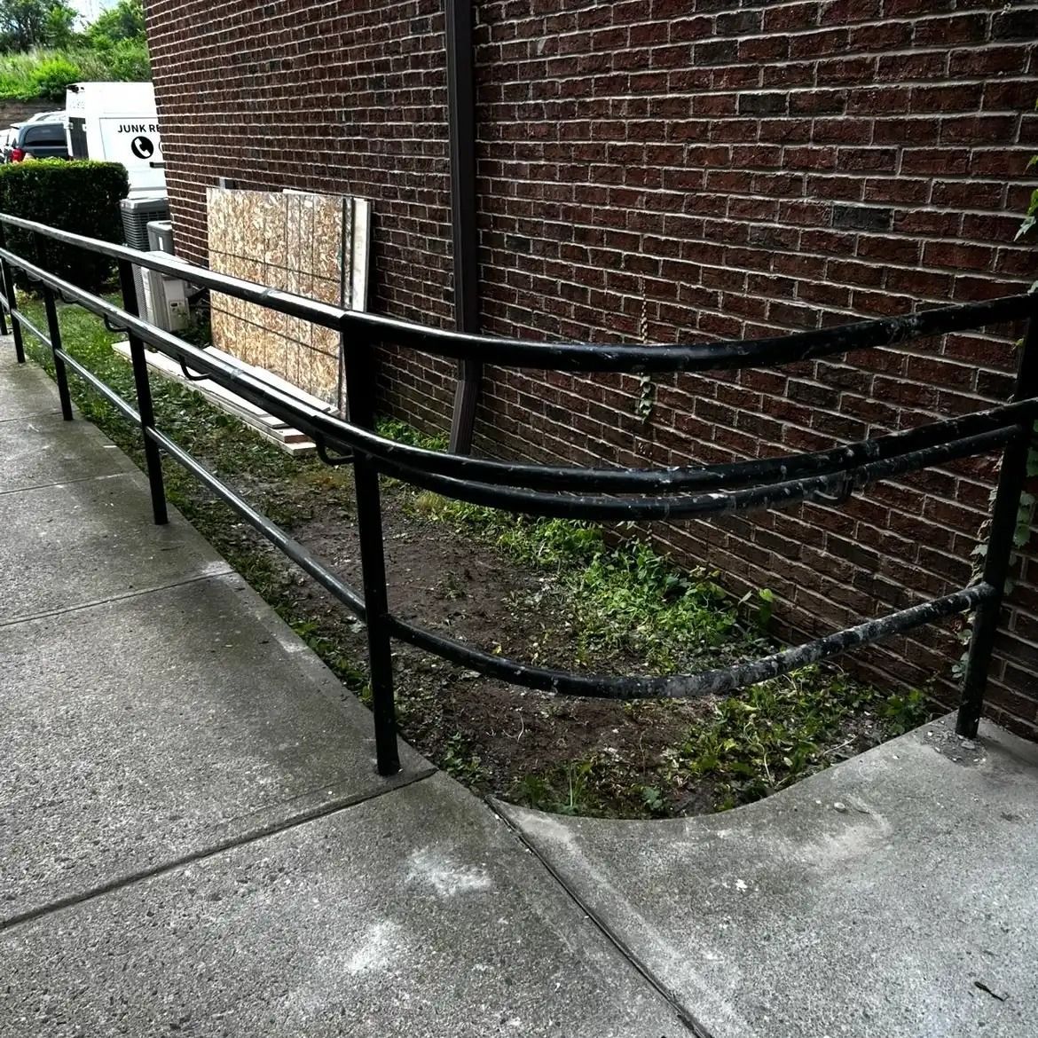 Black metal railings border a concrete ramp beside a brick wall and narrow landscaped strip