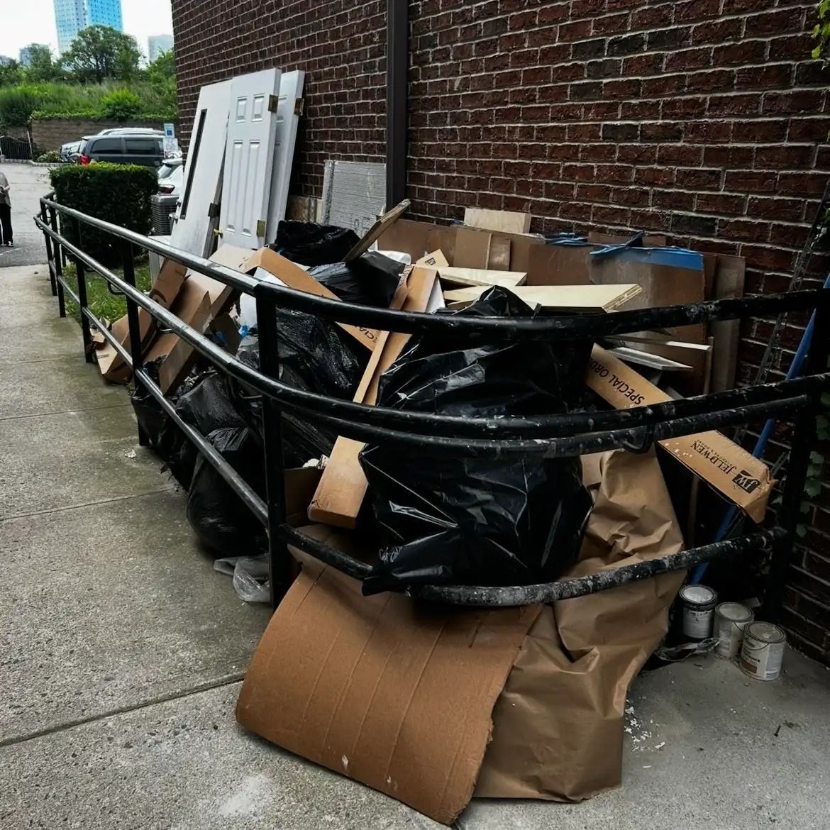 Overflowing recycling cart beside a brick wall, piled with flattened cardboard boxes and black trash bags.