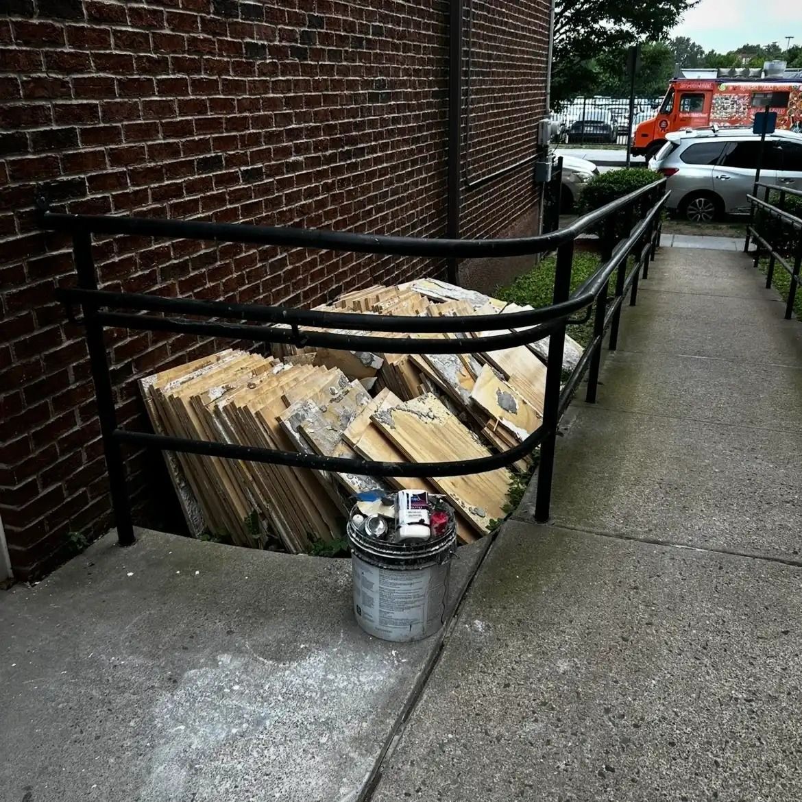 Ramped sidewalk beside brick building with a bent black railing, stacked cardboard, and a bucket on the concrete.