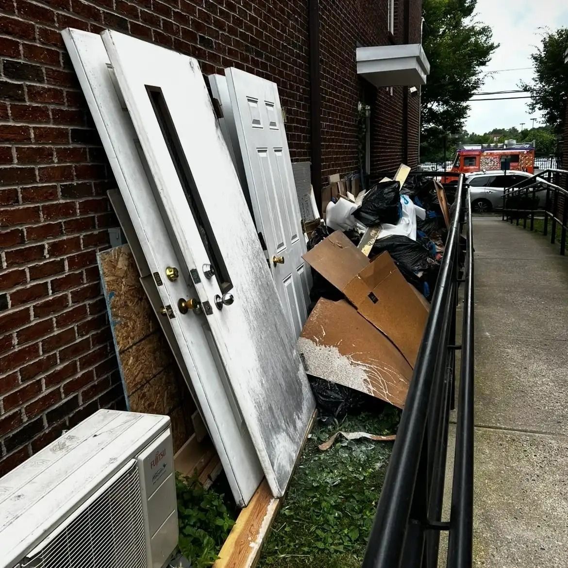 Debris and broken doors piled against a brick building beside a sidewalk and railing