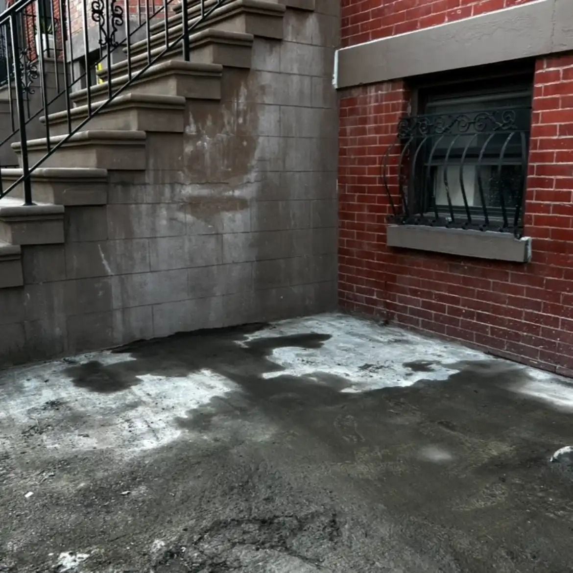 Brick building exterior with concrete steps and a wet, patchy sidewalk beside a barred window