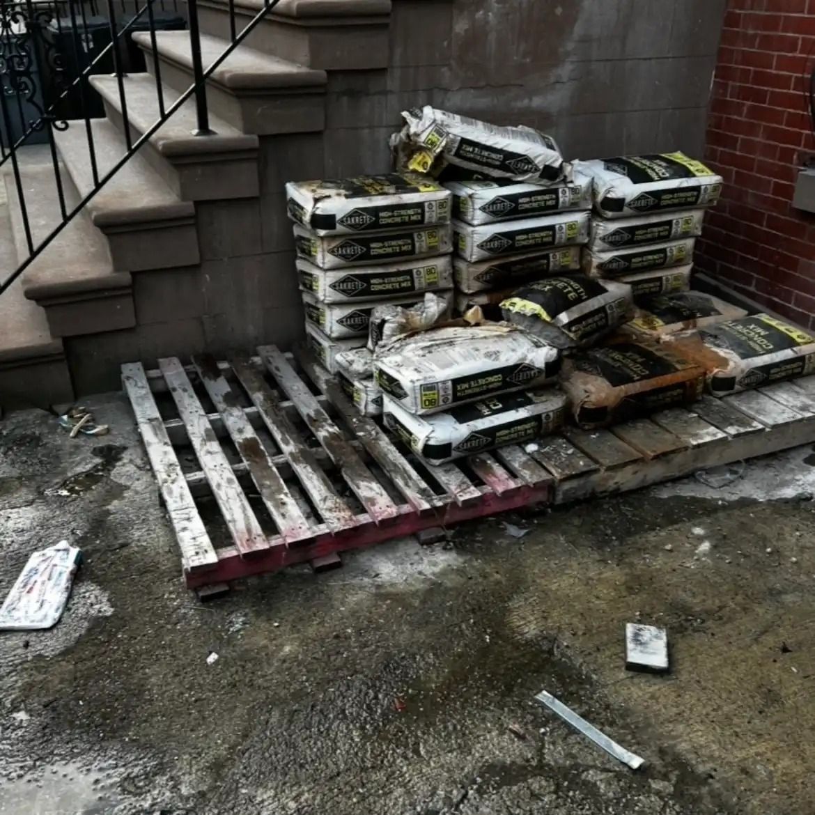 Stacks of bagged material on wooden pallets beside concrete steps and a brick wall.