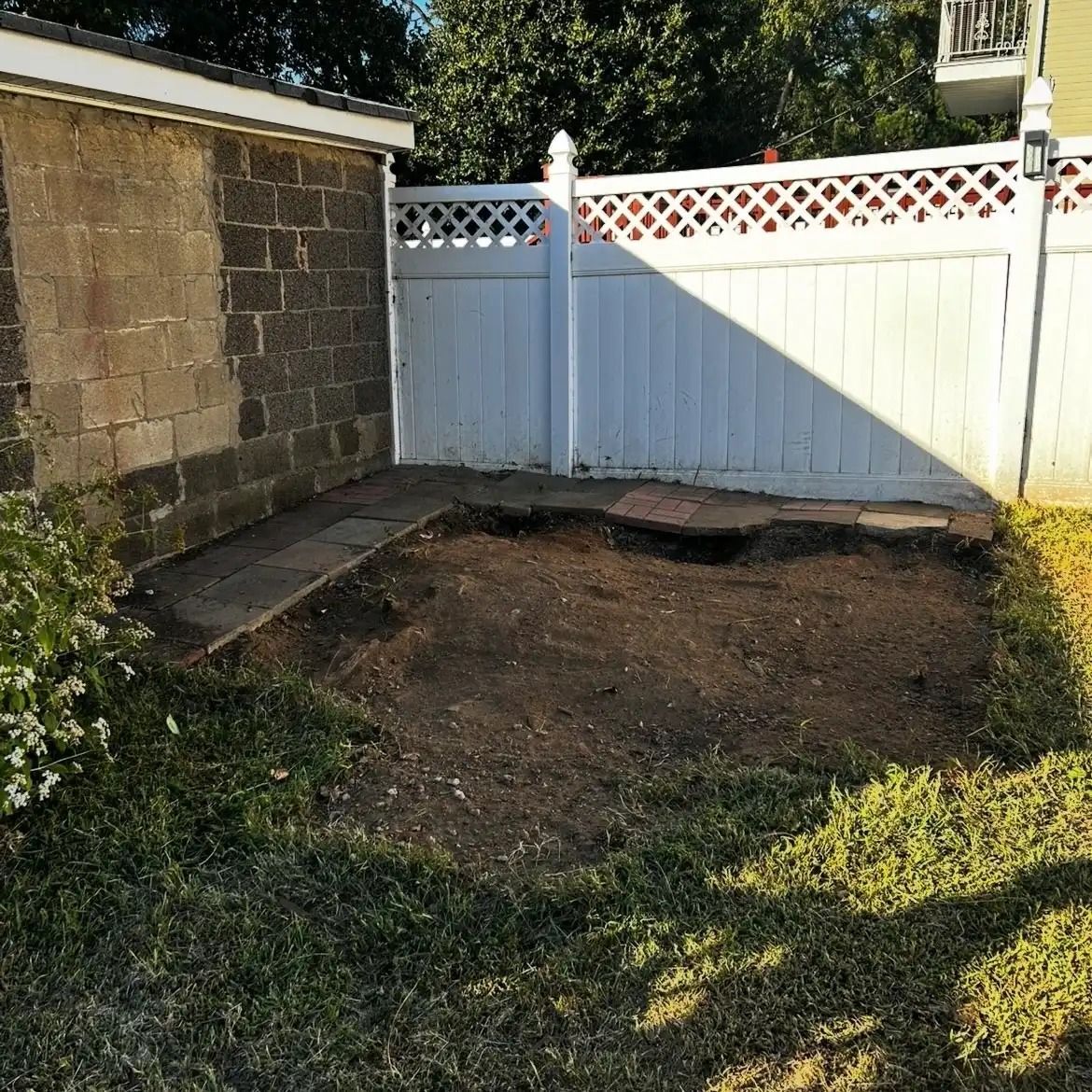 Backyard with bare dirt patch, white fence, stone wall, and green grass in sunlight