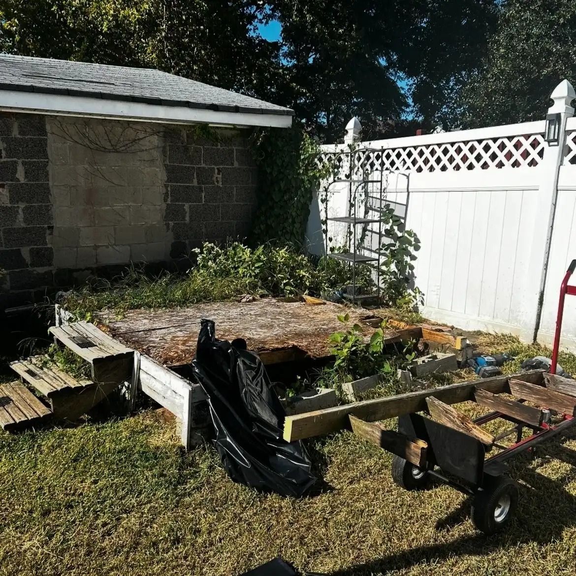 Raised garden bed with black tarp beside a white fence and shed in a sunny backyard