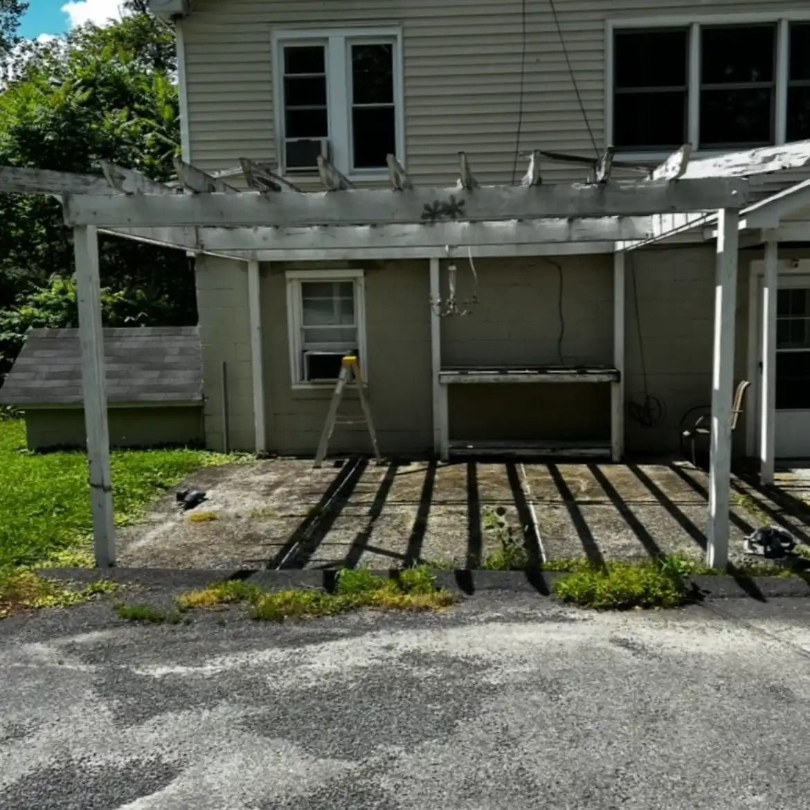 Weathered white pergola over a patio beside a beige house, with a ladder and long shadows on the ground