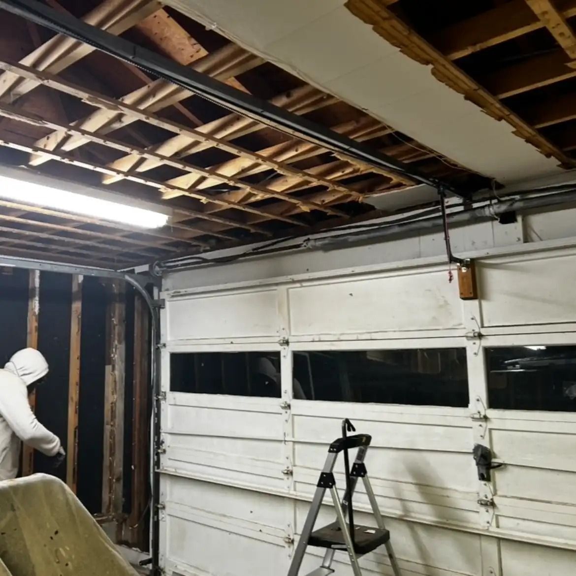 Damaged garage interior with exposed ceiling beams, a worker in a white suit, and a stepladder by a white door