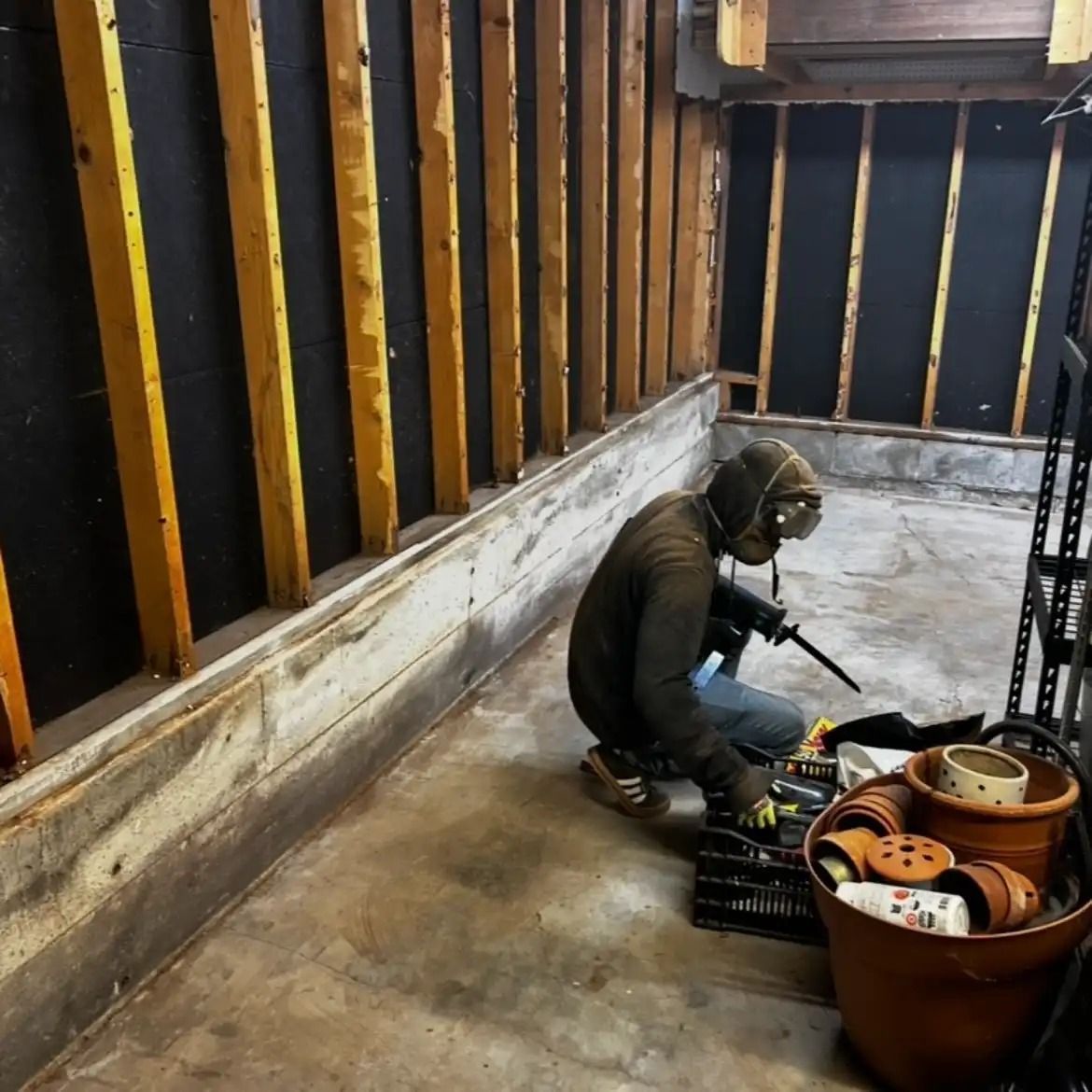 Worker kneeling on a subfloor beside tools and a bucket in a partially framed room.