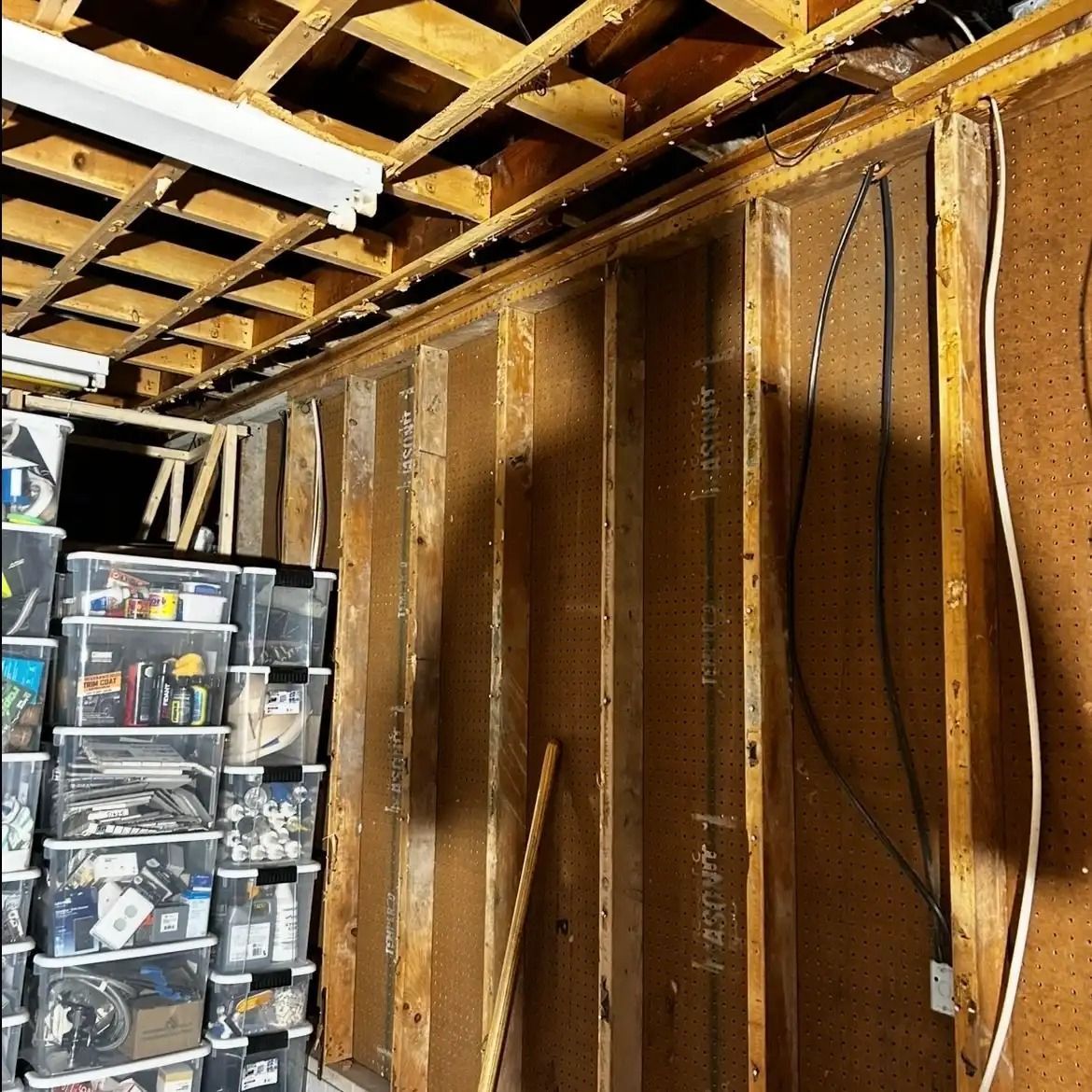 Unfinished basement with exposed ceiling joists, wall studs, hanging wires, and stacked storage bins.