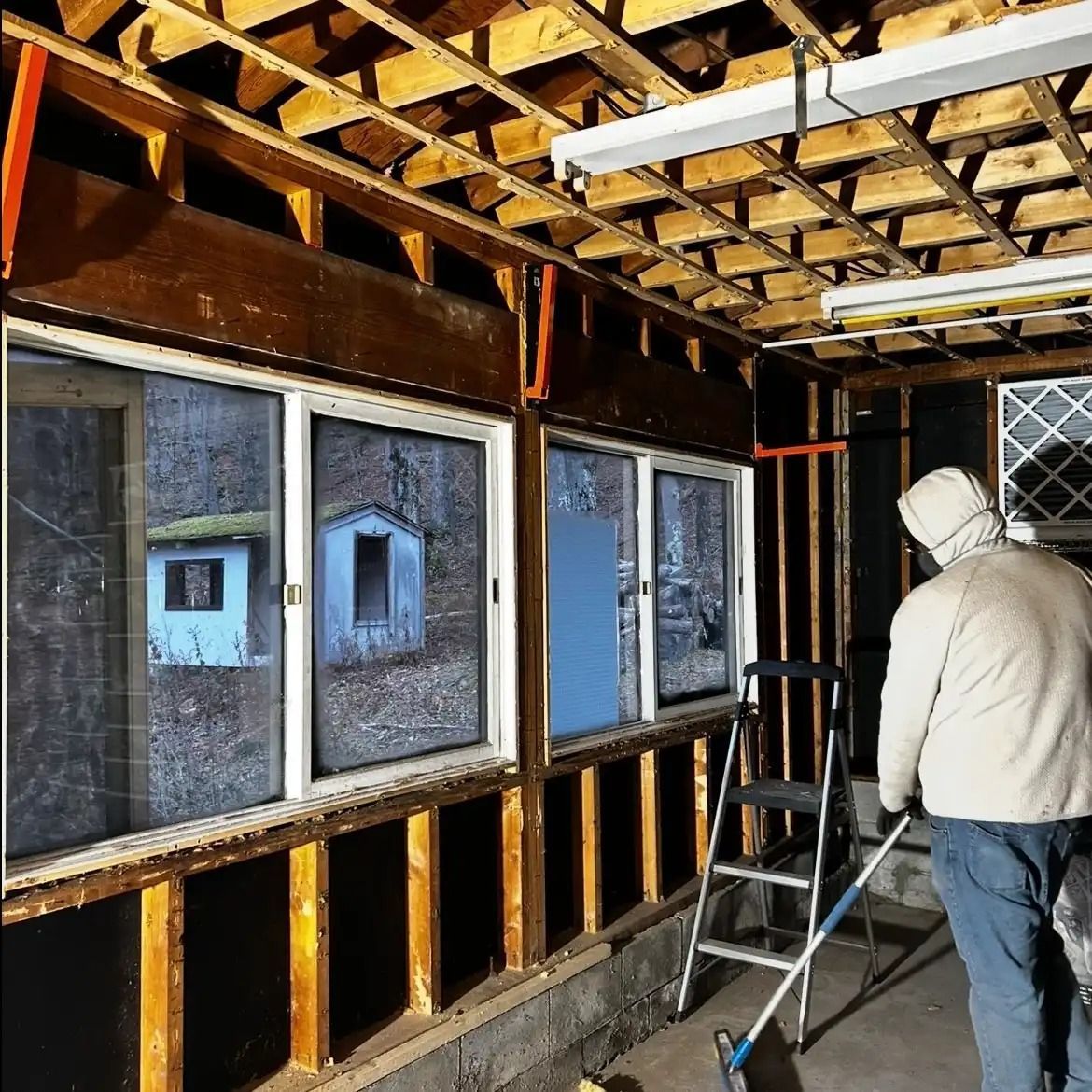 Person in a beige hoodie stands inside a room under renovation, beside a ladder and exposed wall framing.