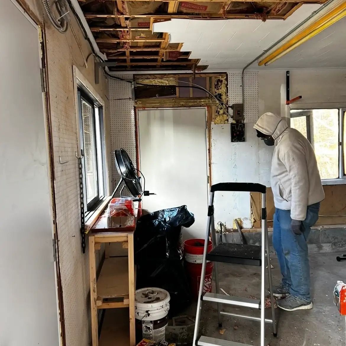 Person in a white hoodie working in a partially demolished room with exposed ceiling and ladder.