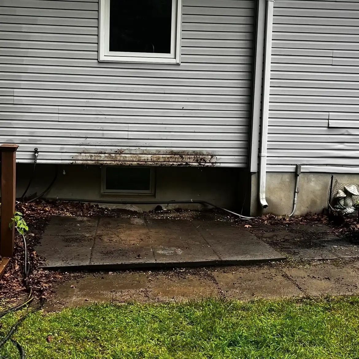 Cracked concrete slab beneath a house with white siding and a small basement window.