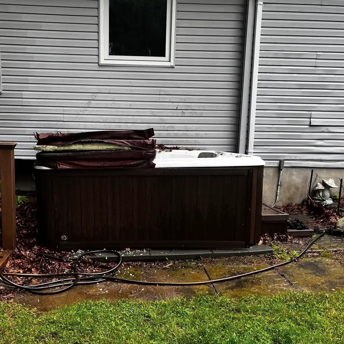 Backyard hot tub against gray house with snow on top and coiled hose on wet grass