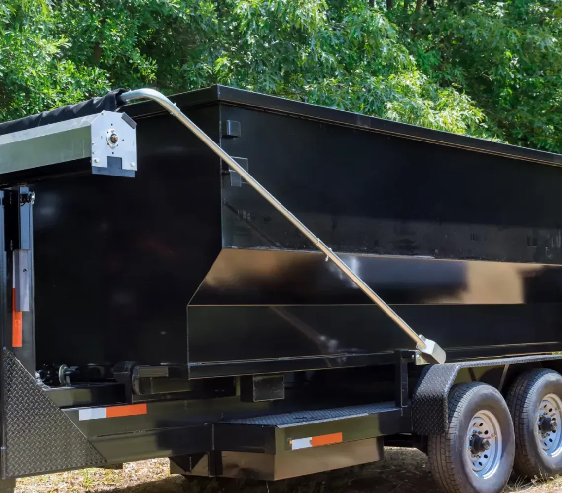 Black dump trailer with tandem axles parked on grass near trees, rear tailgate open.