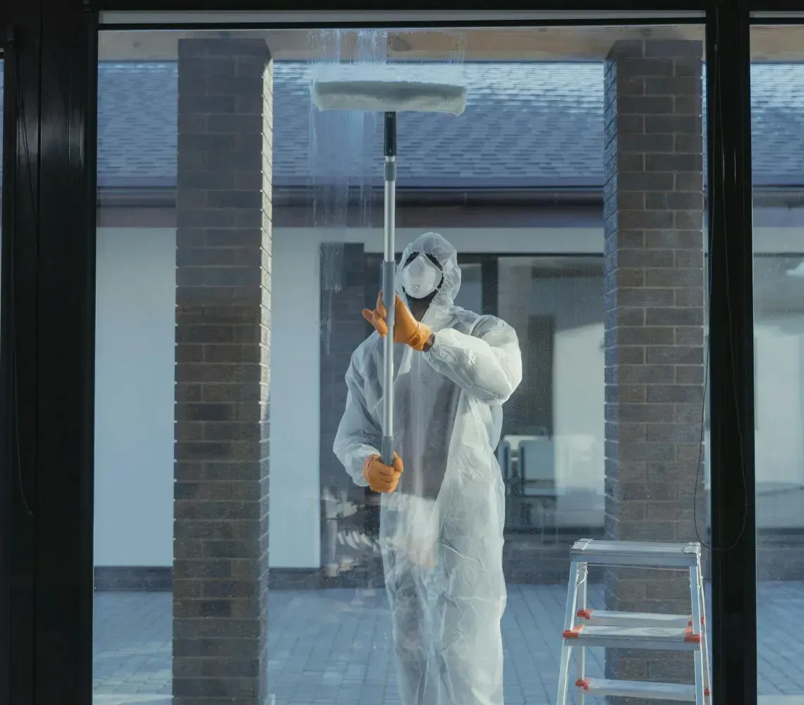 Worker in white coveralls painting a window frame outdoors beside a ladder and blue bucket