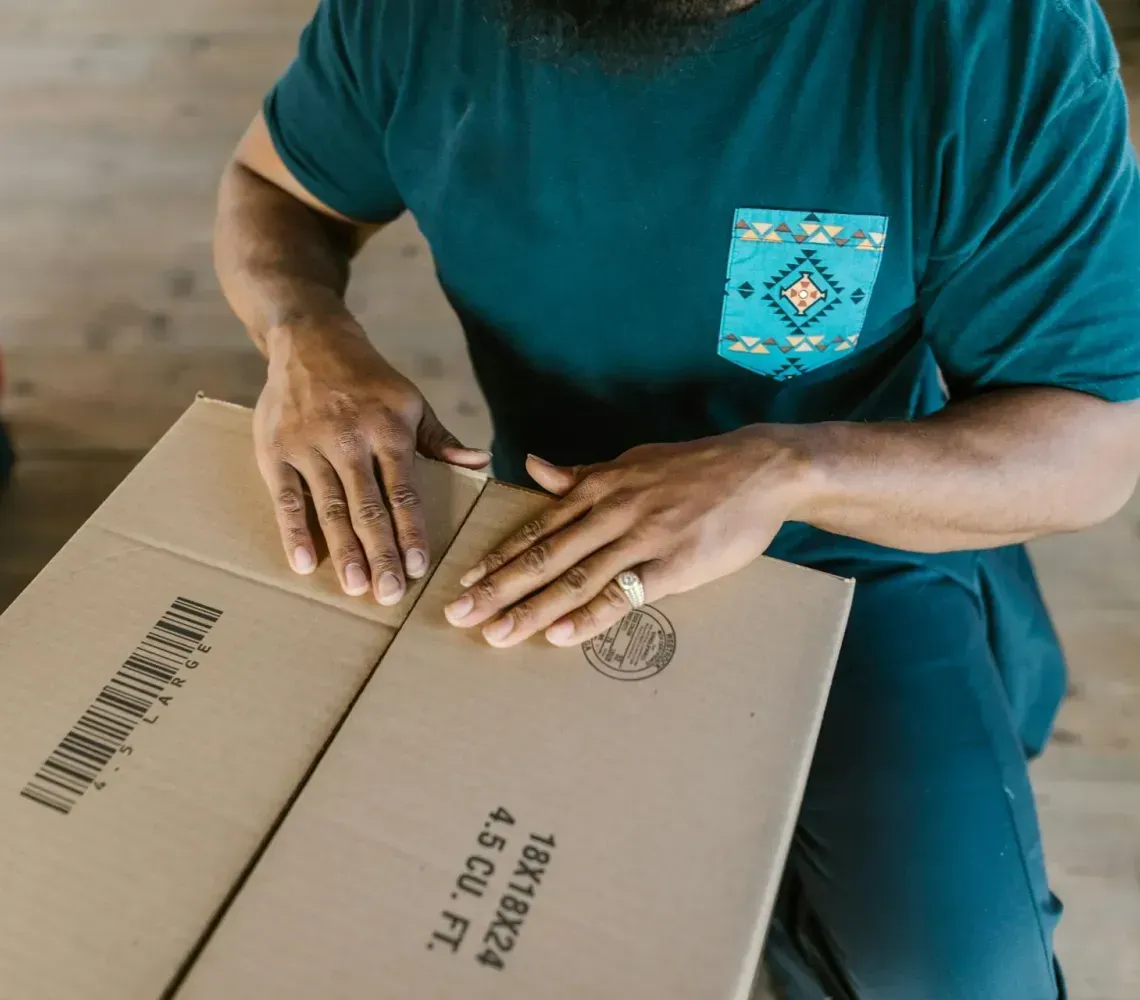 Person in a teal shirt folding a cardboard box on a wooden floor
