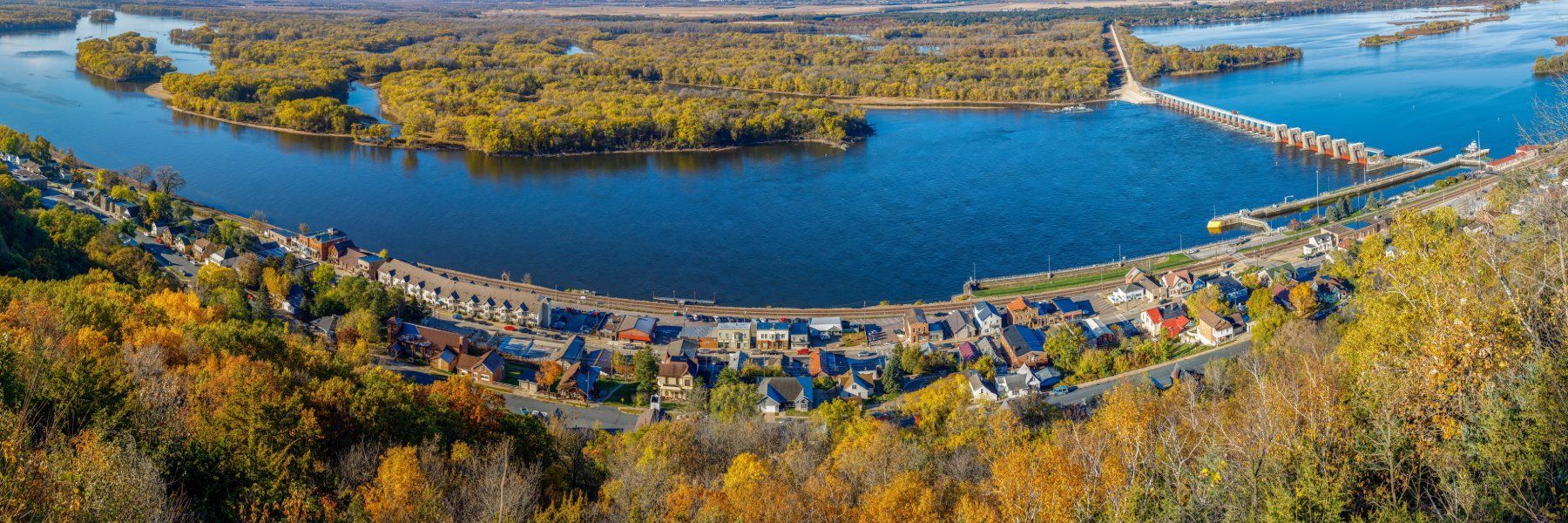Aerial view of a blue river, arched bridge, and tree-lined shore with autumn foliage.