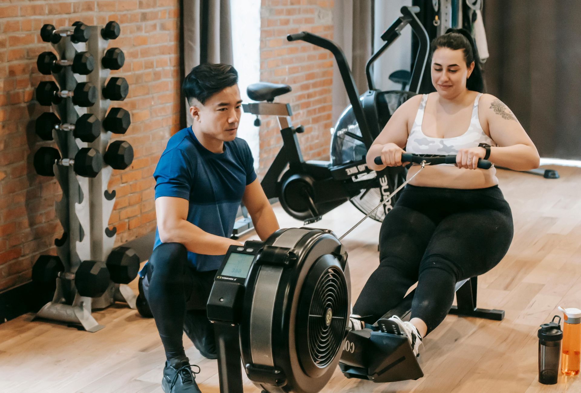 A trainer instructs a person using a rowing machine in a gym with a brick wall and fitness equipment in the background.