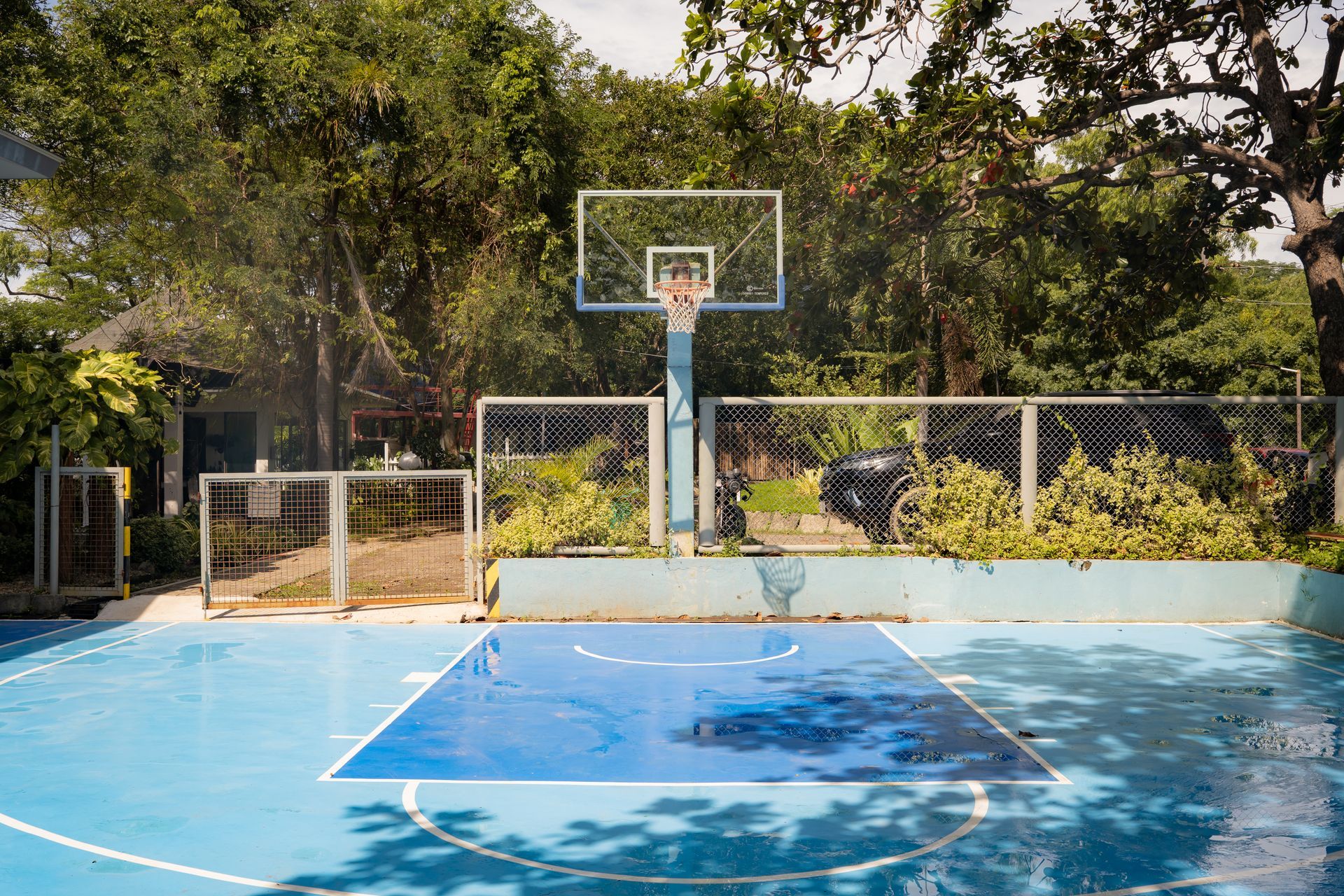 Blue basketball court with a hoop, surrounded by trees and a chain-link fence.