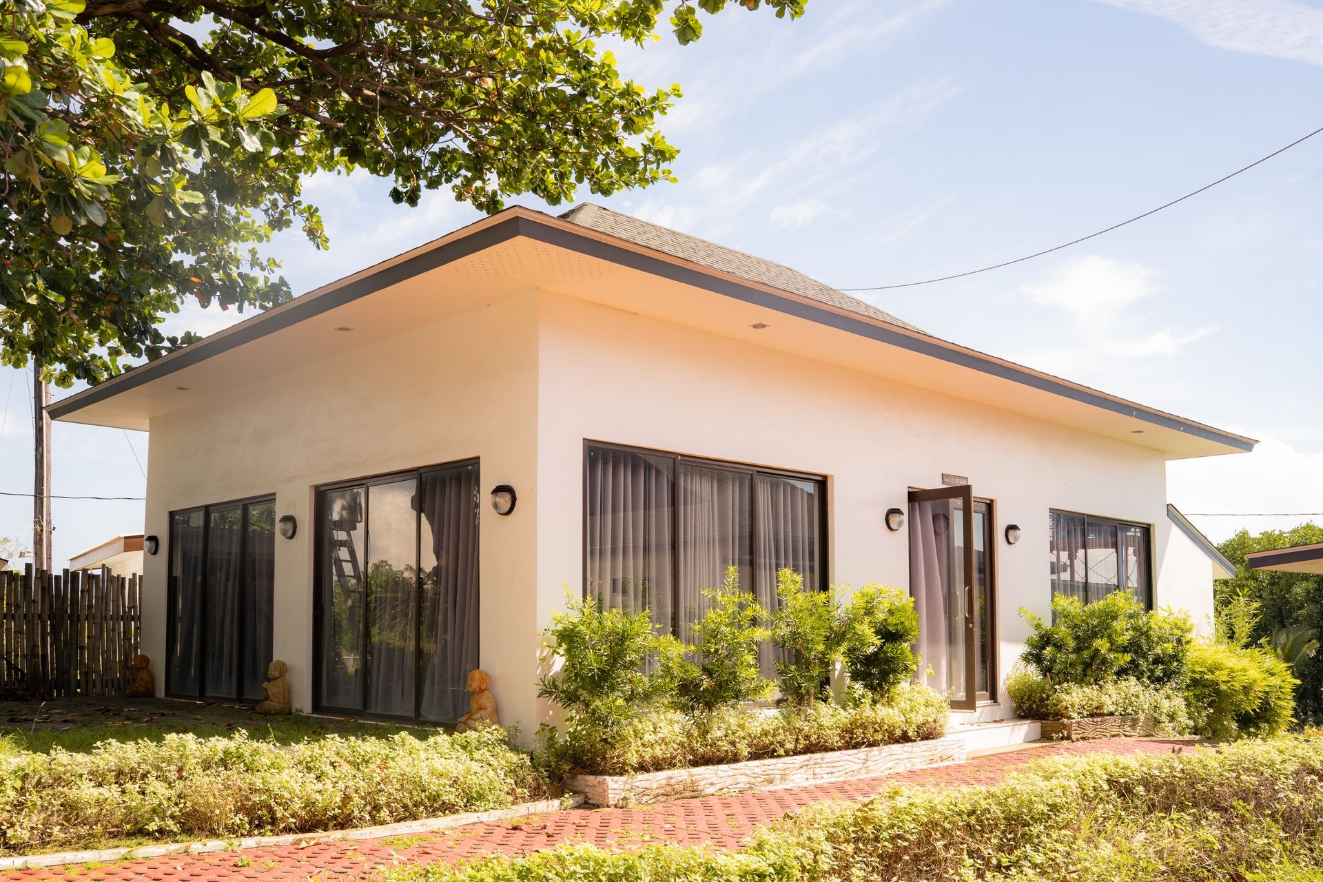 White bungalow with dark-framed windows, surrounded by green shrubs and a brick path, under a blue sky.
