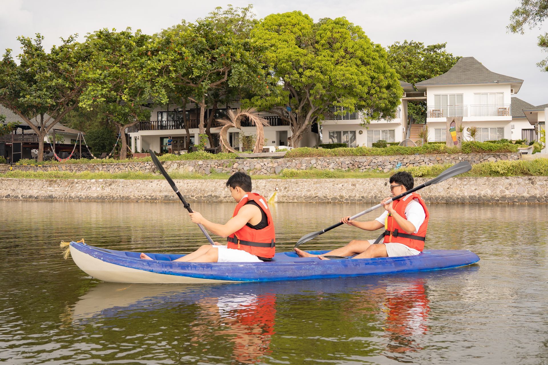 Two people kayaking on water, wearing life vests, in front of a waterfront resort with green trees.