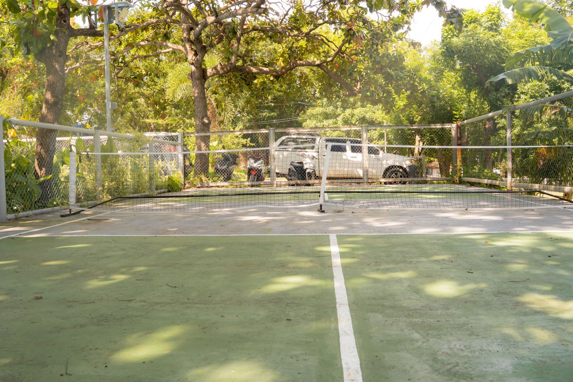 Pickleball court with net, surrounded by a chain-link fence and trees. A white car is visible in the background.