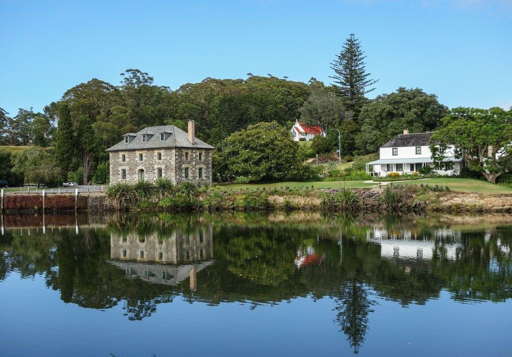 Stone house and white cottage on a calm lakeshore, reflected in still water under a clear blue sky.