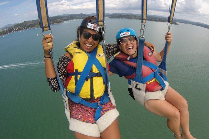 Two people parasail over the water, smiling in blue and pink harnesses.