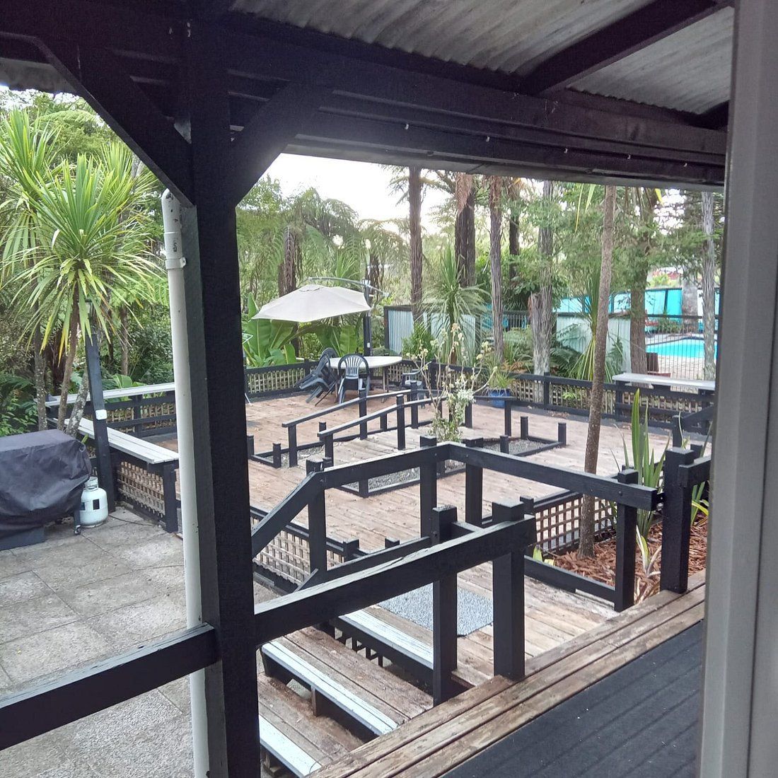 Covered wooden deck overlooking a fenced backyard patio with steps, black railings, and tropical trees.
