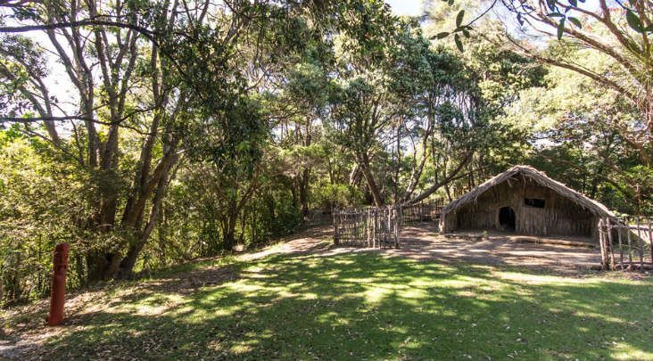 Shaded park clearing with trees, grassy lawn, and a rustic stone shelter with a thatched roof