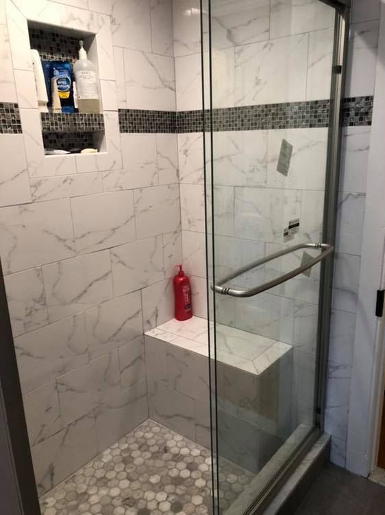 Tiled shower with glass door and built-in bench. White marble-look tiles, mosaic accent. Red shampoo bottle visible.
