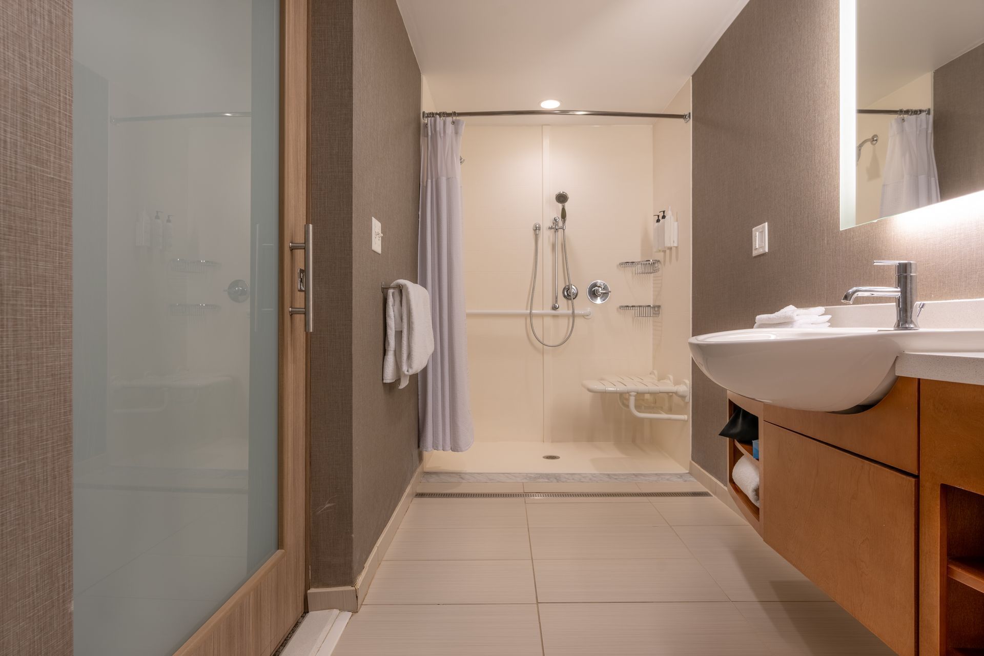 Accessible hotel bathroom with a roll-in shower, sink, and safety bars. Neutral colors.