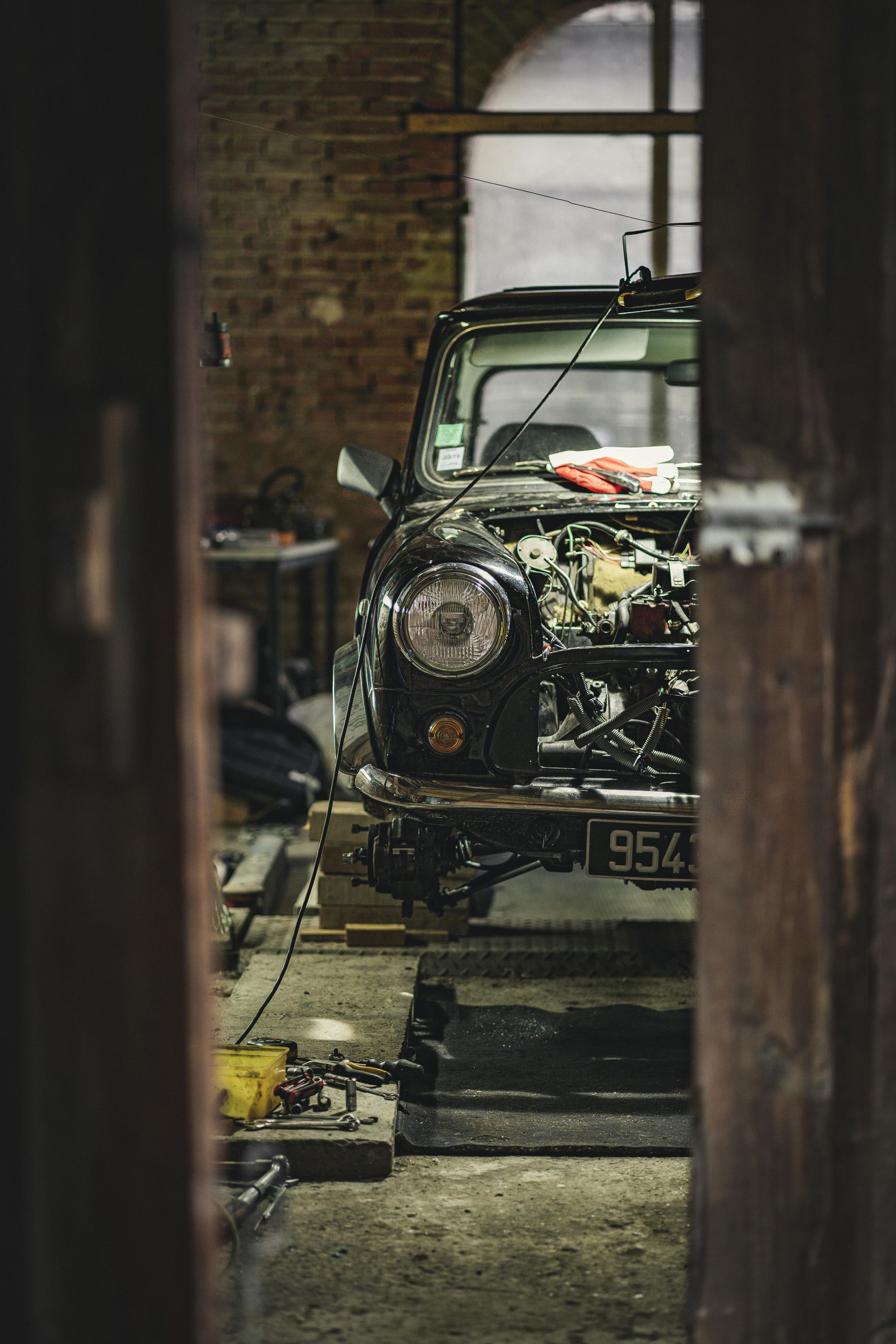 A vintage black car undergoes repair inside a dimly lit brick garage, viewed through a narrow wooden doorway.