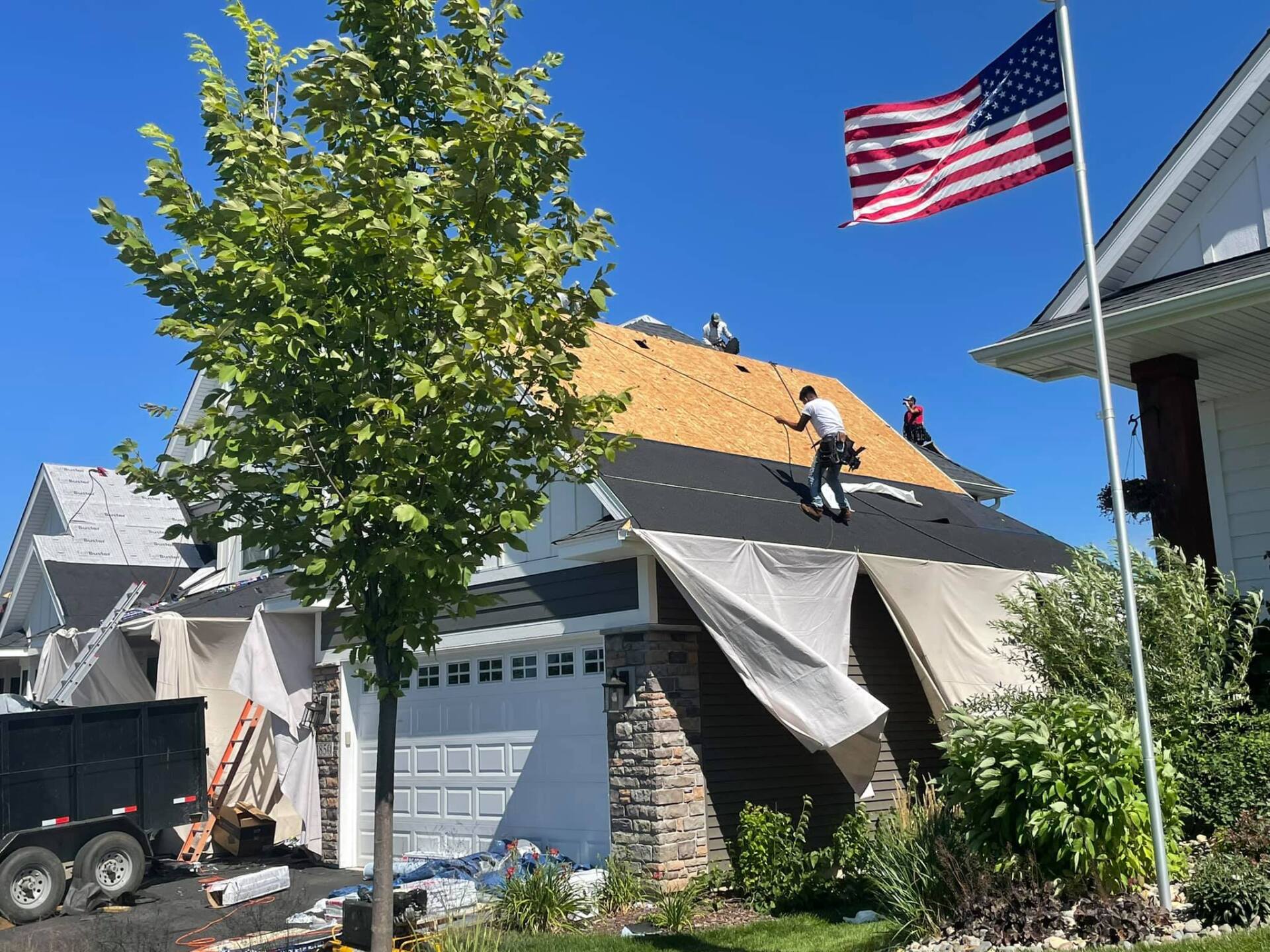 Roofers work on a house with part of the roof exposed. An American flag flies in the background.