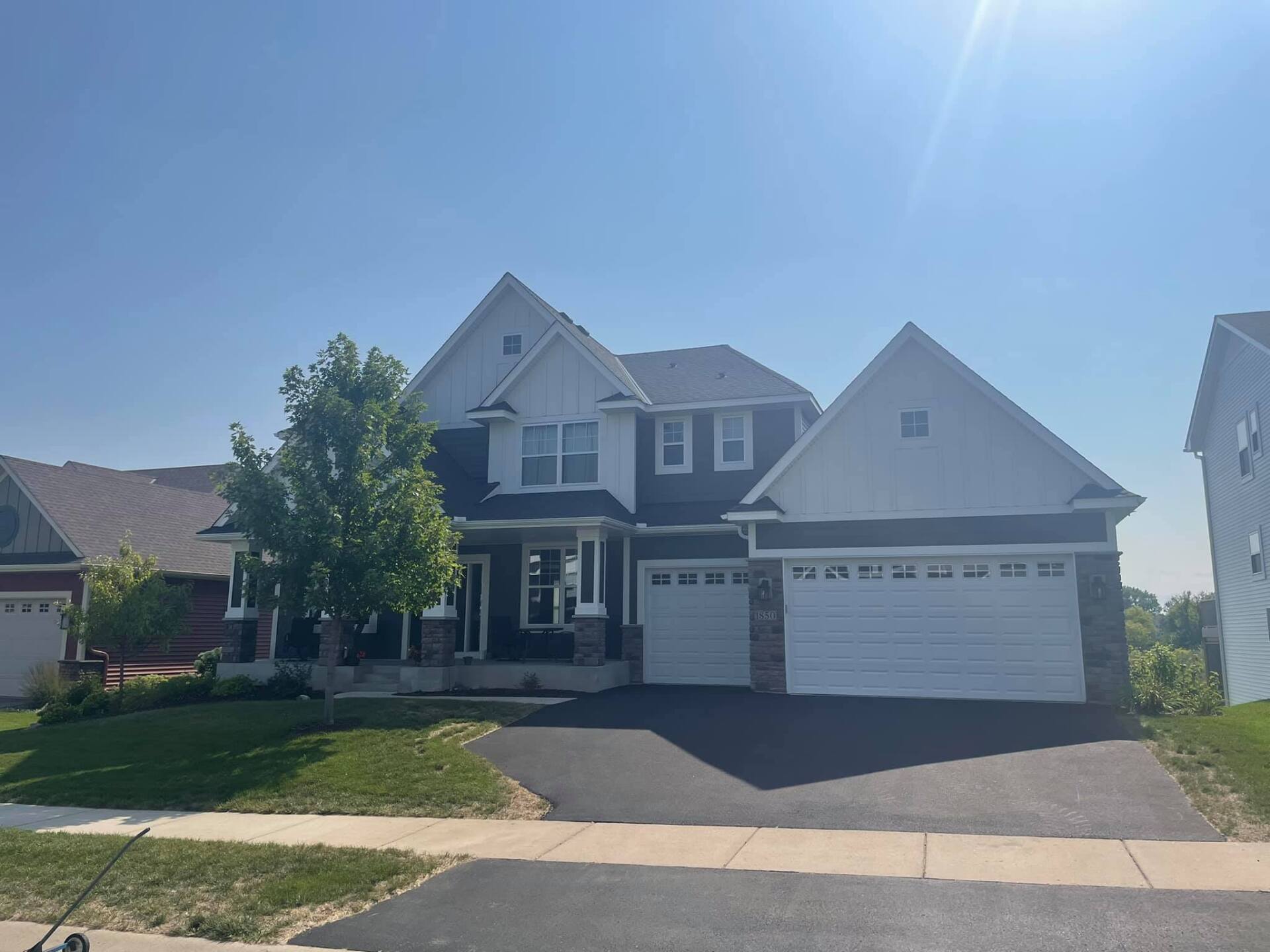 Two-story house with dark gray siding, white trim, and a two-car garage. It has a paved driveway and a well-manicured lawn under a sunny, blue sky.