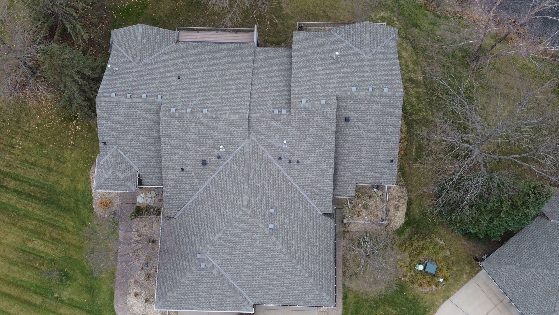 Overhead view of a gray asphalt shingle roof on a house, surrounded by bare trees and grass.