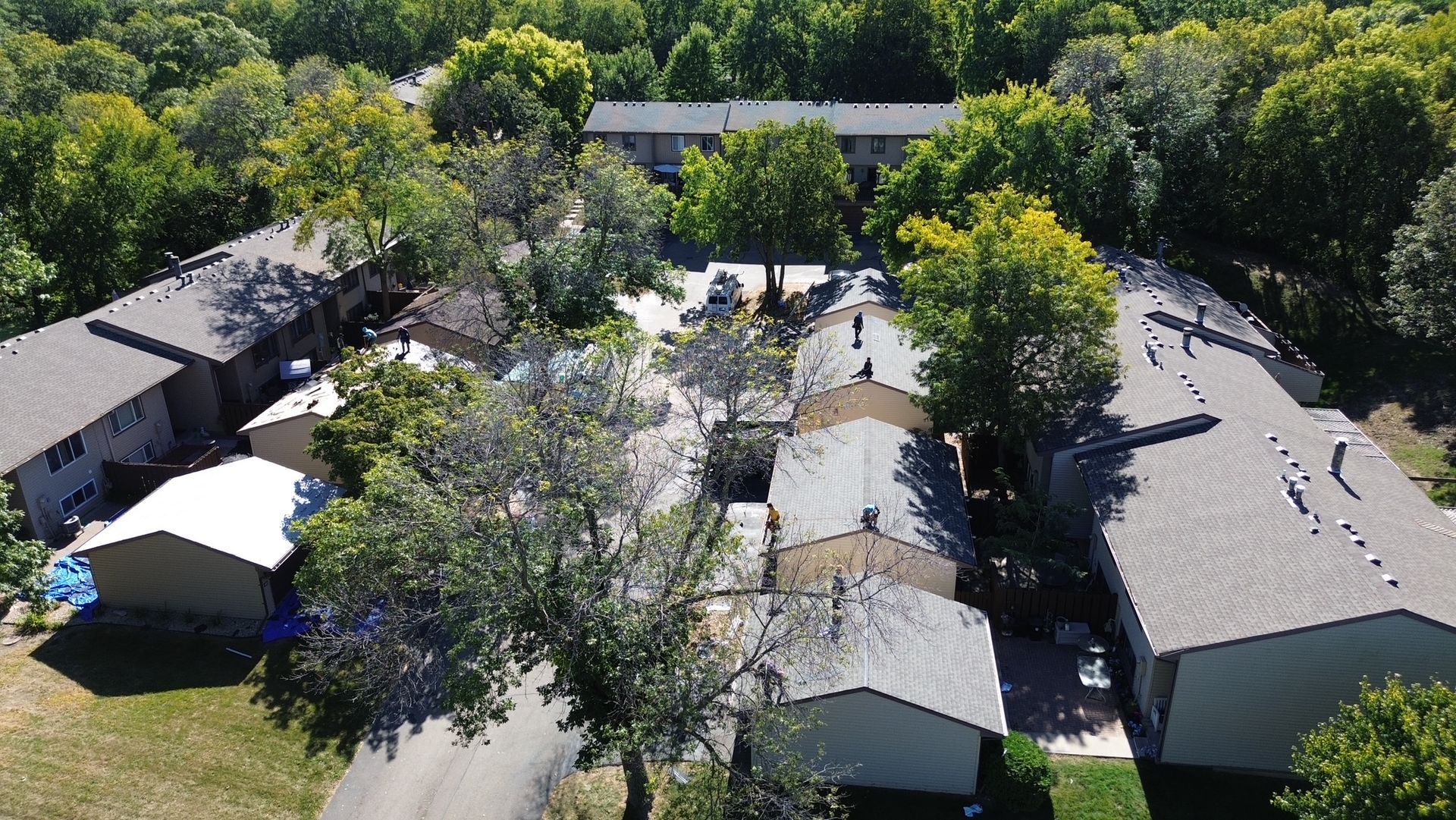 Aerial view of a residential complex with several buildings and surrounding trees. Buildings have gray roofs and are surrounded by greenery.