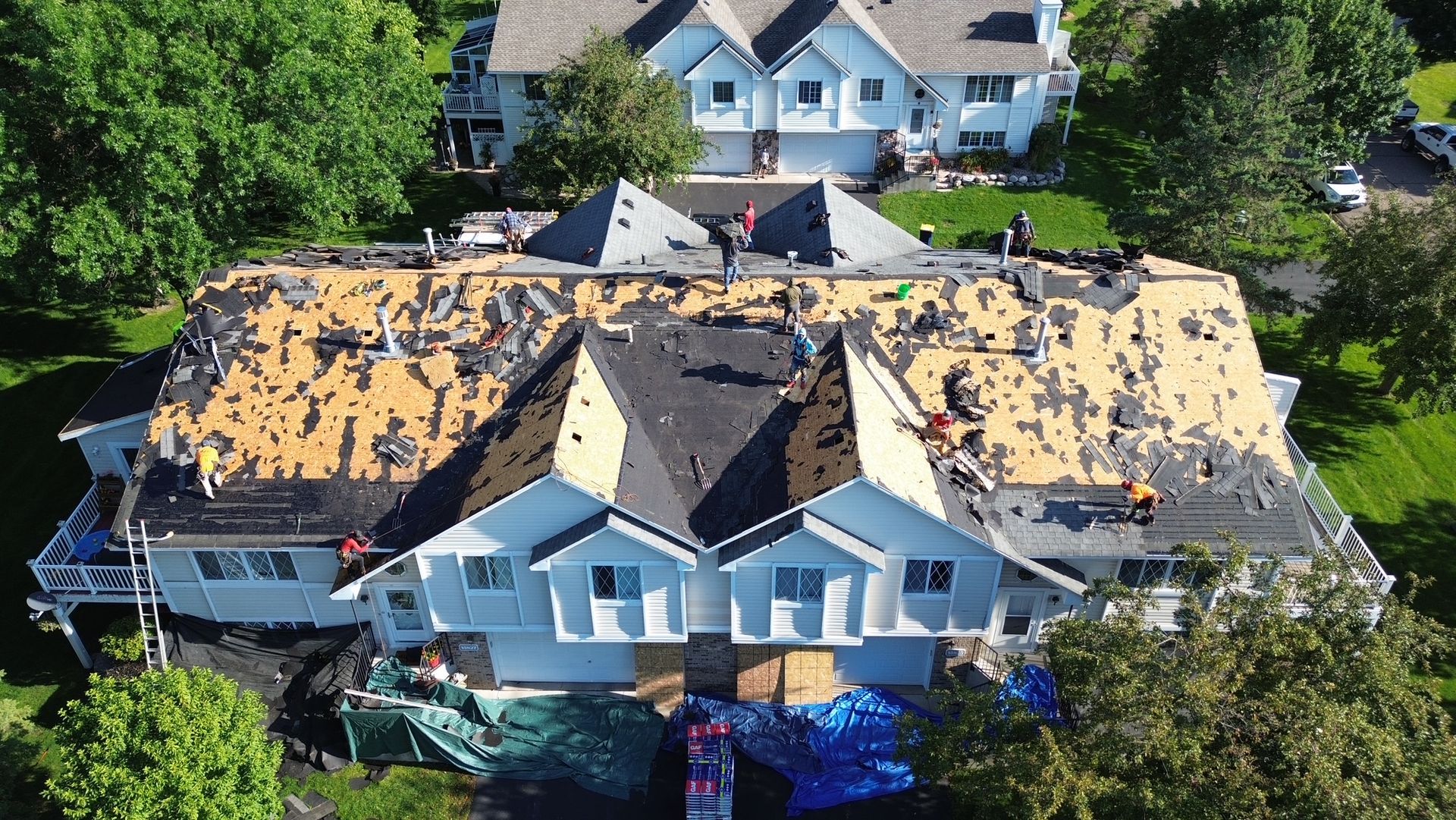 Workers replacing a roof on a light blue two-story house. Black shingles and exposed underlayment are visible. Other houses and green grass are in the background.