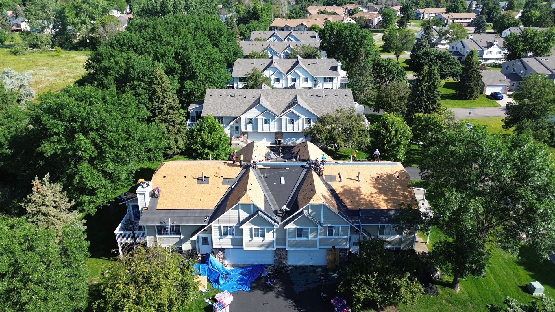 Aerial view of townhouses, some with roofs being replaced, surrounded by trees and green lawns.