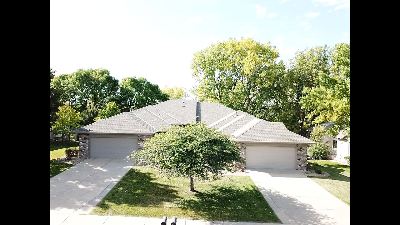 A duplex with gray roofs and tan garages sits in front of lush green trees under a sunny sky.