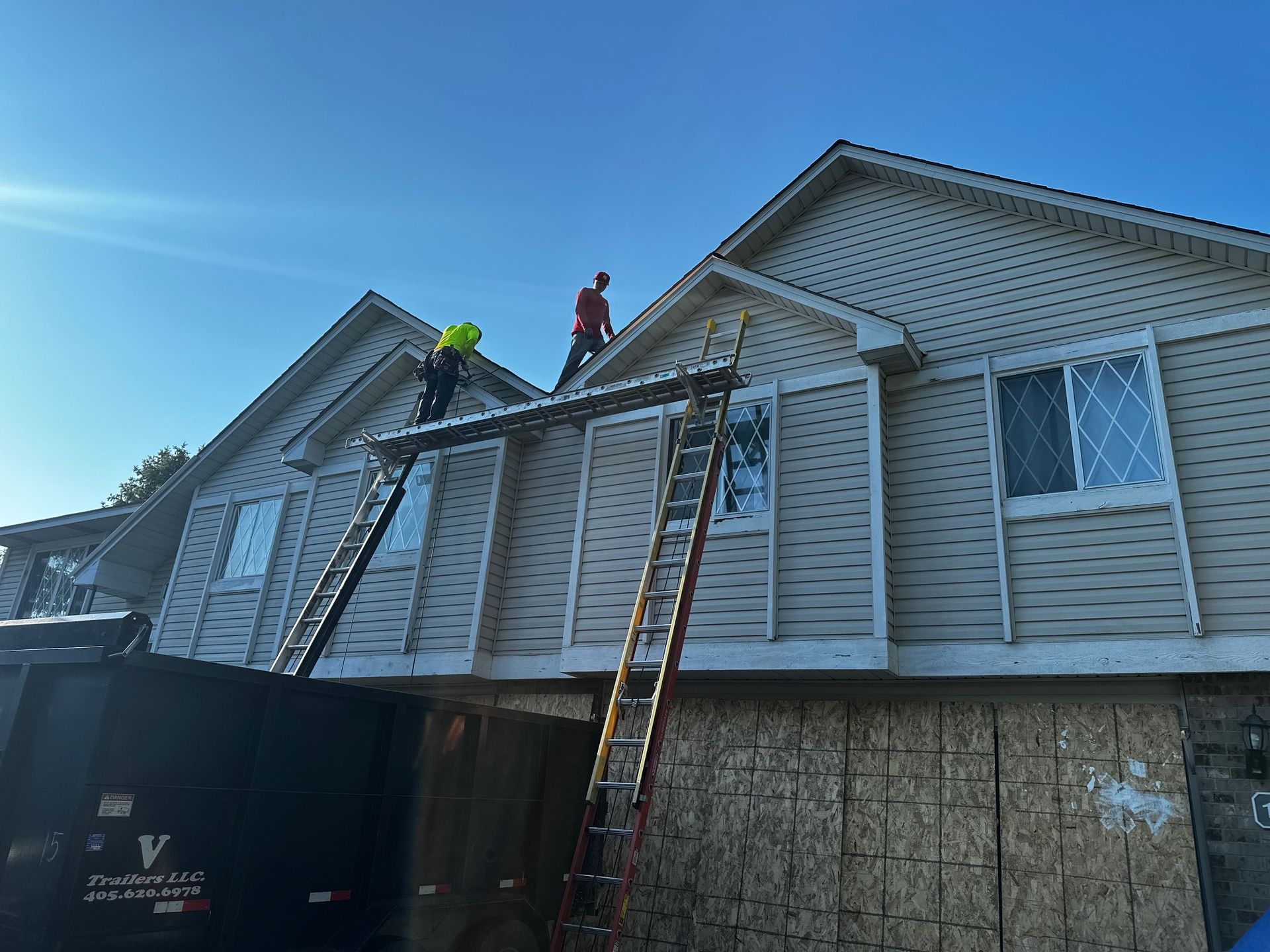 Two construction workers on ladders installing siding on a two-story beige house under a blue sky.