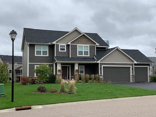 Two-story house with gray siding, a black roof, and a green lawn under a cloudy sky. Includes a lamp post and a two-car garage.
