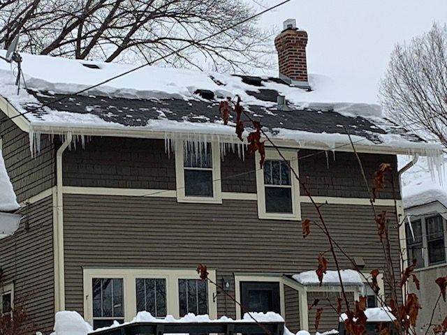 Brown house with snow-covered roof and icicles hanging from the gutters. A brick chimney rises from the roof.