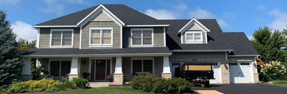A two-story house with a dark roof and gray siding, a large front porch, and a two-car garage.  A dark-colored vehicle is parked in the garage.
