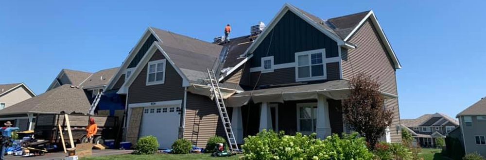 Roofing work in progress on a two-story house with dark brown siding and a white garage door. A ladder is propped against the roof, where a worker is visible.