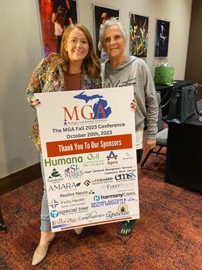 Two women hold a colorful sign at an indoor event, smiling beside a table.