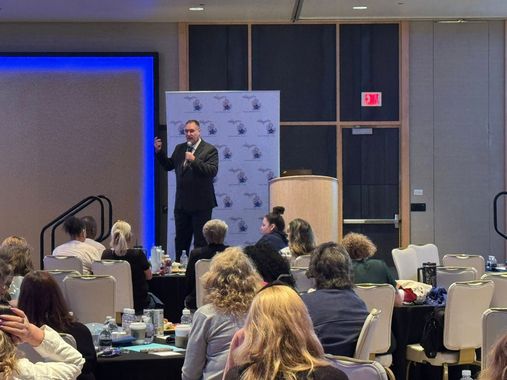 Speaker presenting at a conference podium while audience sits at round tables in a hotel ballroom