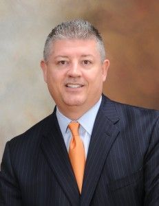 Portrait of a man in a dark pinstripe suit with an orange tie against a warm studio backdrop