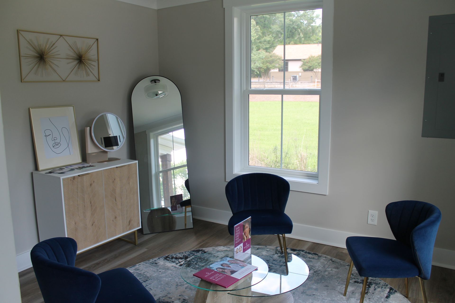 Modern living room with blue chairs, glass coffee table, cabinet, mirror, and large window view